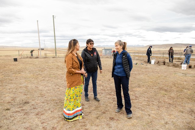 Three women facing each other, talking in dry field. In the background, people are working with shovels.