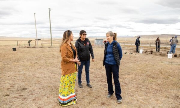 Three women facing each other, talking in dry field. In the background, people are working with shovels.