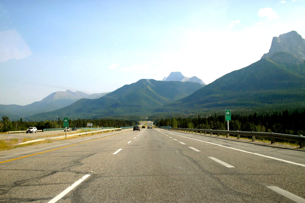Blue sky and green mountains rise above a highway that stretches ahead of the viewer.